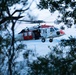A U.S. Navy search and rescue helicopter, identifiable by its white and red color scheme and 'NAVY' markings, is captured in low-level flight framed through tree branches near a body of water. The aircraft appears to be a Sikorsky MH-60S Knighthawk or similar variant, with its side door open and a crew member visible inside. The image is taken at dusk or dawn, giving the scene a cool blue tone.