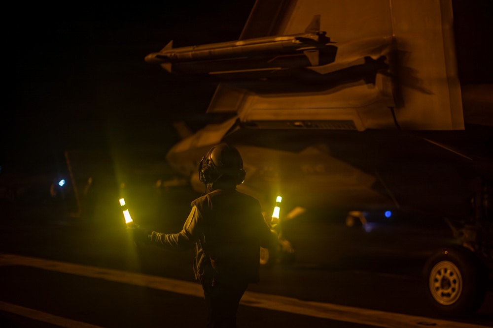 A flight deck crew member is conducting night operations on what appears to be an aircraft carrier flight deck, using illuminated marshalling wands to guide aircraft. The scene is dominated by low-light conditions with warm amber/yellow tones from deck lighting. A military aircraft wing with an air-to-air missile mounted is visible overhead.