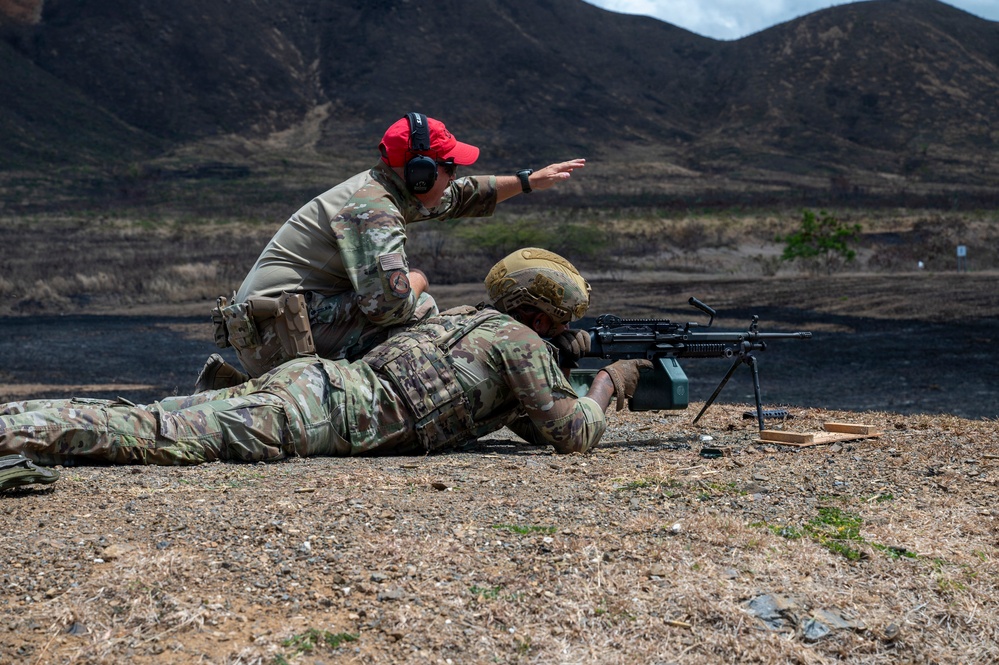 Two military personnel are at an outdoor firing range, with one soldier in the prone position operating a light machine gun mounted on a bipod, while an instructor kneels behind and gestures toward the target area. The setting appears to be an arid, mountainous training range. This appears to be a live-fire training exercise with an instructor providing guidance.