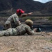 Two military personnel are at an outdoor firing range, with one soldier in the prone position operating a light machine gun mounted on a bipod, while an instructor kneels behind and gestures toward the target area. The setting appears to be an arid, mountainous training range. This appears to be a live-fire training exercise with an instructor providing guidance.