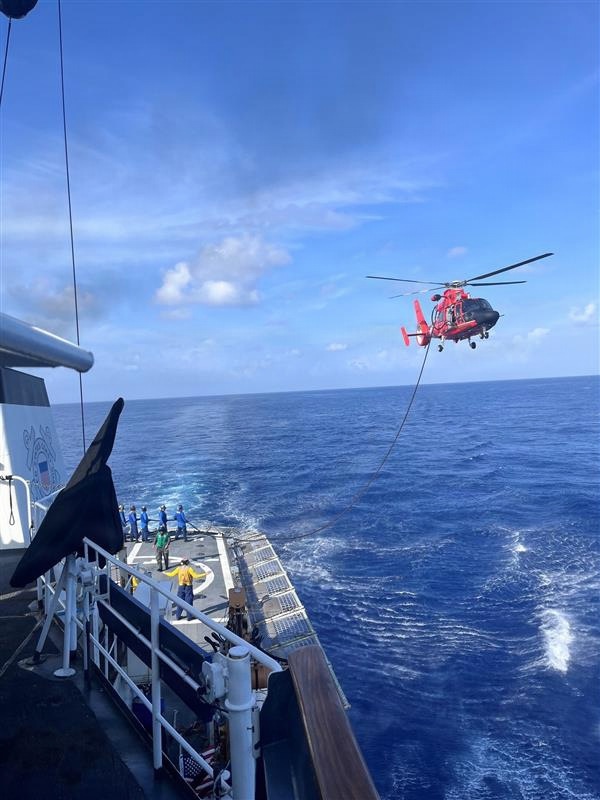 A red and black helicopter, identifiable as a U.S. Coast Guard MH-65 Dolphin, hovers off the stern of a Coast Guard cutter over open ocean while connected by a hoist cable to the ship's flight deck. Crew members in various colored vests and uniforms are positioned on the flight deck, with one individual in a yellow vest appearing to signal with arms raised. This appears to be a hoist training exercise or active rescue/transfer operation at sea.