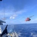 A red and black helicopter, identifiable as a U.S. Coast Guard MH-65 Dolphin, hovers off the stern of a Coast Guard cutter over open ocean while connected by a hoist cable to the ship's flight deck. Crew members in various colored vests and uniforms are positioned on the flight deck, with one individual in a yellow vest appearing to signal with arms raised. This appears to be a hoist training exercise or active rescue/transfer operation at sea.
