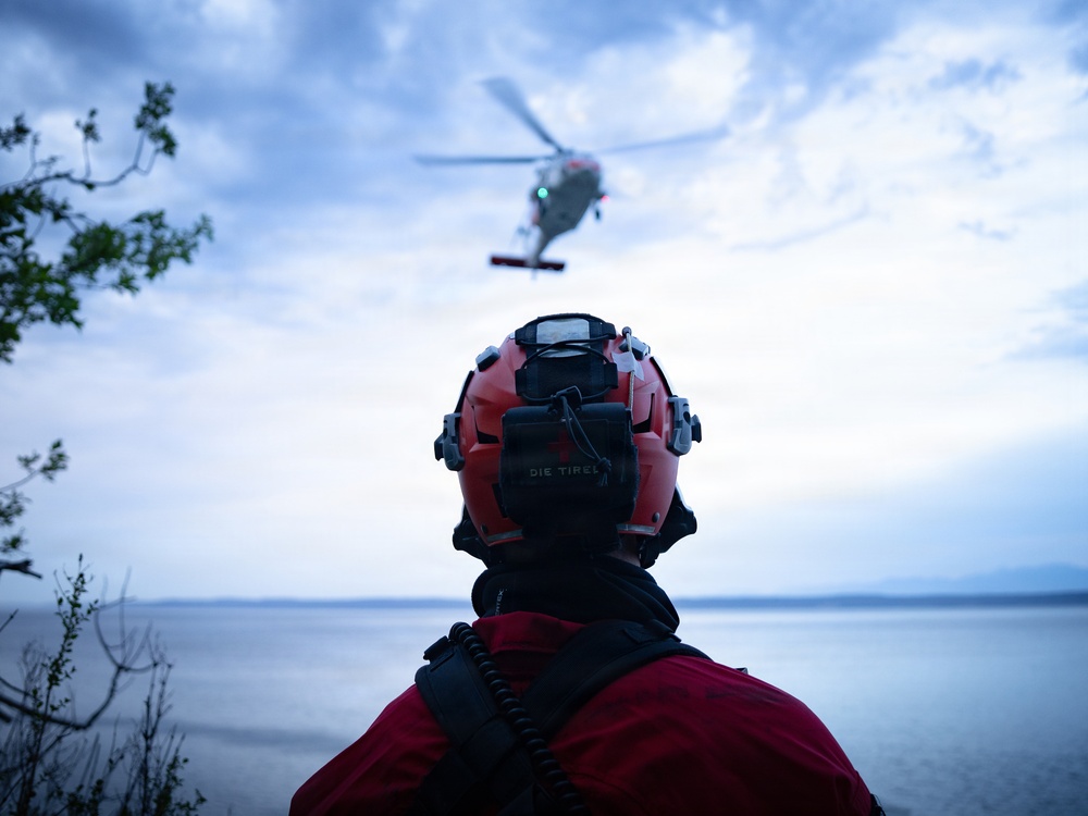 A rescue or emergency personnel wearing a red helmet with communications equipment observes a helicopter hovering over a body of water during overcast conditions. The individual is viewed from behind, standing on an elevated rocky or vegetated shoreline. The scene strongly suggests an active search and rescue or emergency response operation.