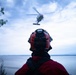 A rescue or emergency personnel wearing a red helmet with communications equipment observes a helicopter hovering over a body of water during overcast conditions. The individual is viewed from behind, standing on an elevated rocky or vegetated shoreline. The scene strongly suggests an active search and rescue or emergency response operation.
