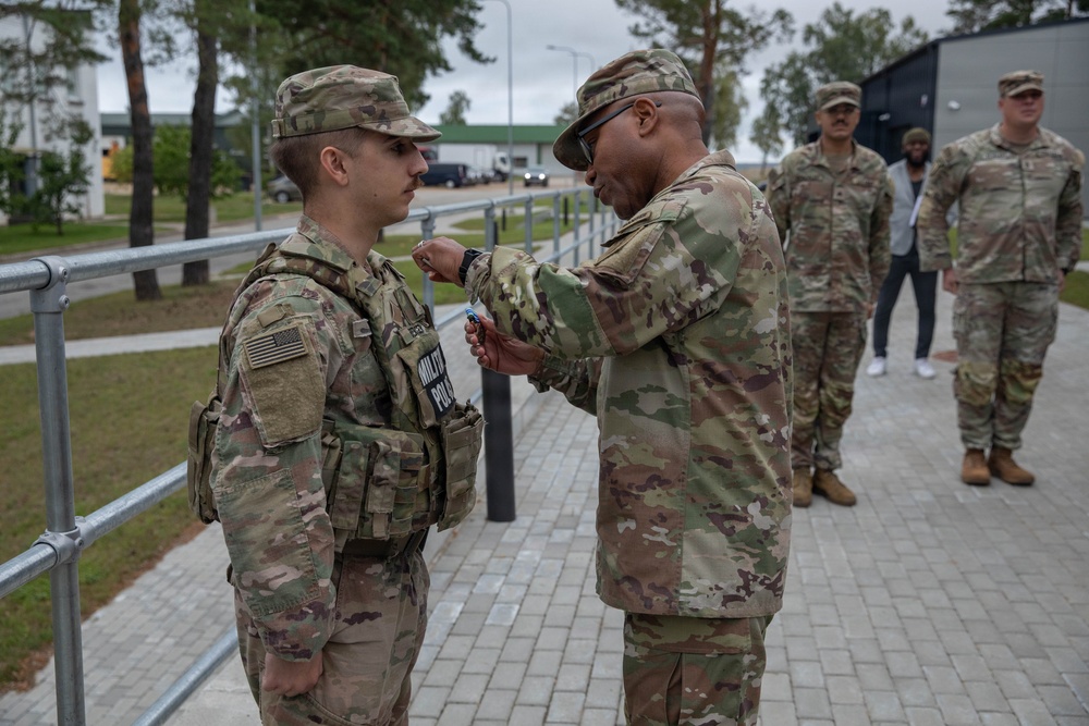 A senior U.S. Army soldier appears to be pinning a medal or award onto a junior soldier wearing a Military Police vest and full combat gear. The ceremony is taking place outdoors on a paved walkway, with several other uniformed soldiers observing in the background. The setting appears to be a military installation, likely in Europe based on the architecture and paving style.
