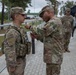 A senior U.S. Army soldier appears to be pinning a medal or award onto a junior soldier wearing a Military Police vest and full combat gear. The ceremony is taking place outdoors on a paved walkway, with several other uniformed soldiers observing in the background. The setting appears to be a military installation, likely in Europe based on the architecture and paving style.