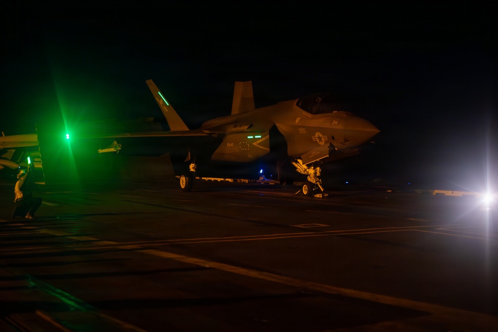 A military jet aircraft, identifiable as an F-35 variant, is photographed on what appears to be an aircraft carrier flight deck during nighttime operations. The aircraft is illuminated by green navigation lights and a bright white light source on the right side of the frame. At least one ground crew member is visible near the aircraft.