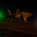 A military jet aircraft, identifiable as an F-35 variant, is photographed on what appears to be an aircraft carrier flight deck during nighttime operations. The aircraft is illuminated by green navigation lights and a bright white light source on the right side of the frame. At least one ground crew member is visible near the aircraft.