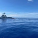A U.S. Coast Guard cutter is positioned on open ocean alongside a small orange inflatable rescue boat (RIB) that appears to be transiting toward a sailboat in the distance. The scene suggests an active maritime interdiction, rescue, or boarding operation in calm, deep blue ocean waters. Three distinct vessels are visible, indicating a coordinated Coast Guard operation.