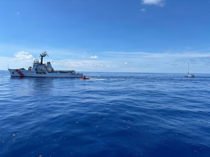 A U.S. Coast Guard cutter is positioned on open ocean alongside a small orange inflatable rescue boat (RIB) that appears to be transiting toward a sailboat in the distance. The scene suggests an active maritime interdiction, rescue, or boarding operation in calm, deep blue ocean waters. Three distinct vessels are visible, indicating a coordinated Coast Guard operation.