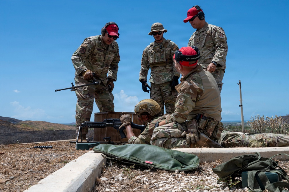 A group of military personnel in camouflage uniforms are conducting a weapons training exercise at an outdoor firing range. Several individuals wearing red hearing protection caps are observing and instructing while one person is in a prone shooting position behind a rifle. The setting appears to be an elevated outdoor range with arid terrain and a clear blue sky in the background.