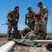 A group of military personnel in camouflage uniforms are conducting a weapons training exercise at an outdoor firing range. Several individuals wearing red hearing protection caps are observing and instructing while one person is in a prone shooting position behind a rifle. The setting appears to be an elevated outdoor range with arid terrain and a clear blue sky in the background.