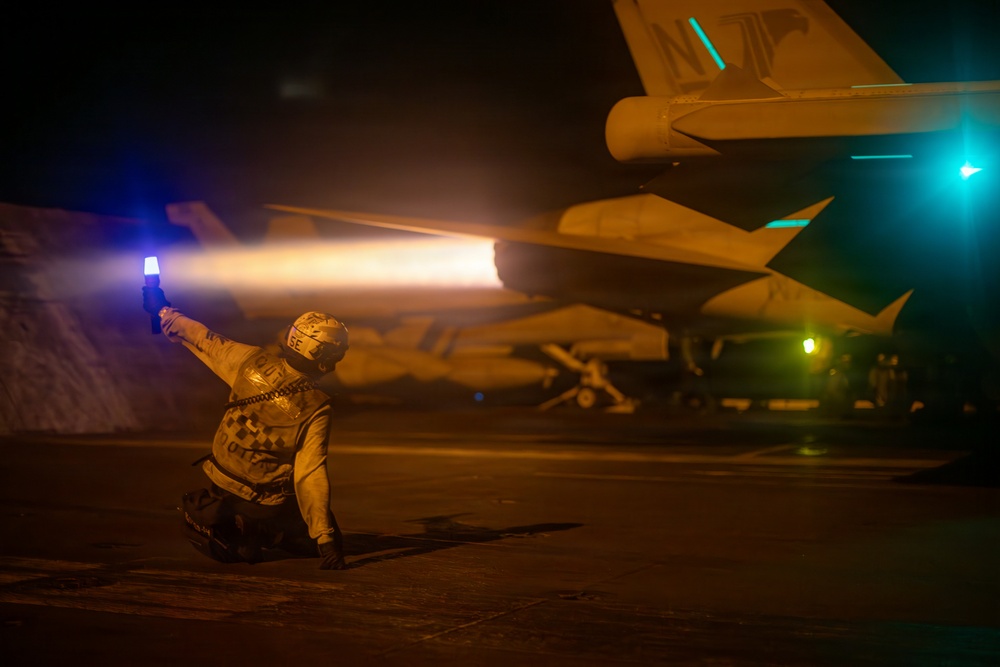 A flight deck crew member kneels on an aircraft carrier flight deck at night, holding a blue illuminated wand while directing a military jet aircraft. The jet's afterburner is visibly ignited, producing a bright orange flame plume. The scene is illuminated by a combination of the afterburner glow, the blue wand light, and green navigation/deck lights in the background.