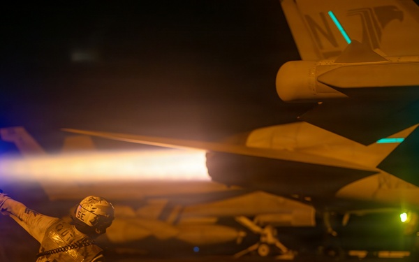 A flight deck crew member kneels on an aircraft carrier flight deck at night, holding a blue illuminated wand while directing a military jet aircraft. The jet's afterburner is visibly ignited, producing a bright orange flame plume. The scene is illuminated by a combination of the afterburner glow, the blue wand light, and green navigation/deck lights in the background.
