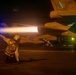 A flight deck crew member kneels on an aircraft carrier flight deck at night, holding a blue illuminated wand while directing a military jet aircraft. The jet's afterburner is visibly ignited, producing a bright orange flame plume. The scene is illuminated by a combination of the afterburner glow, the blue wand light, and green navigation/deck lights in the background.