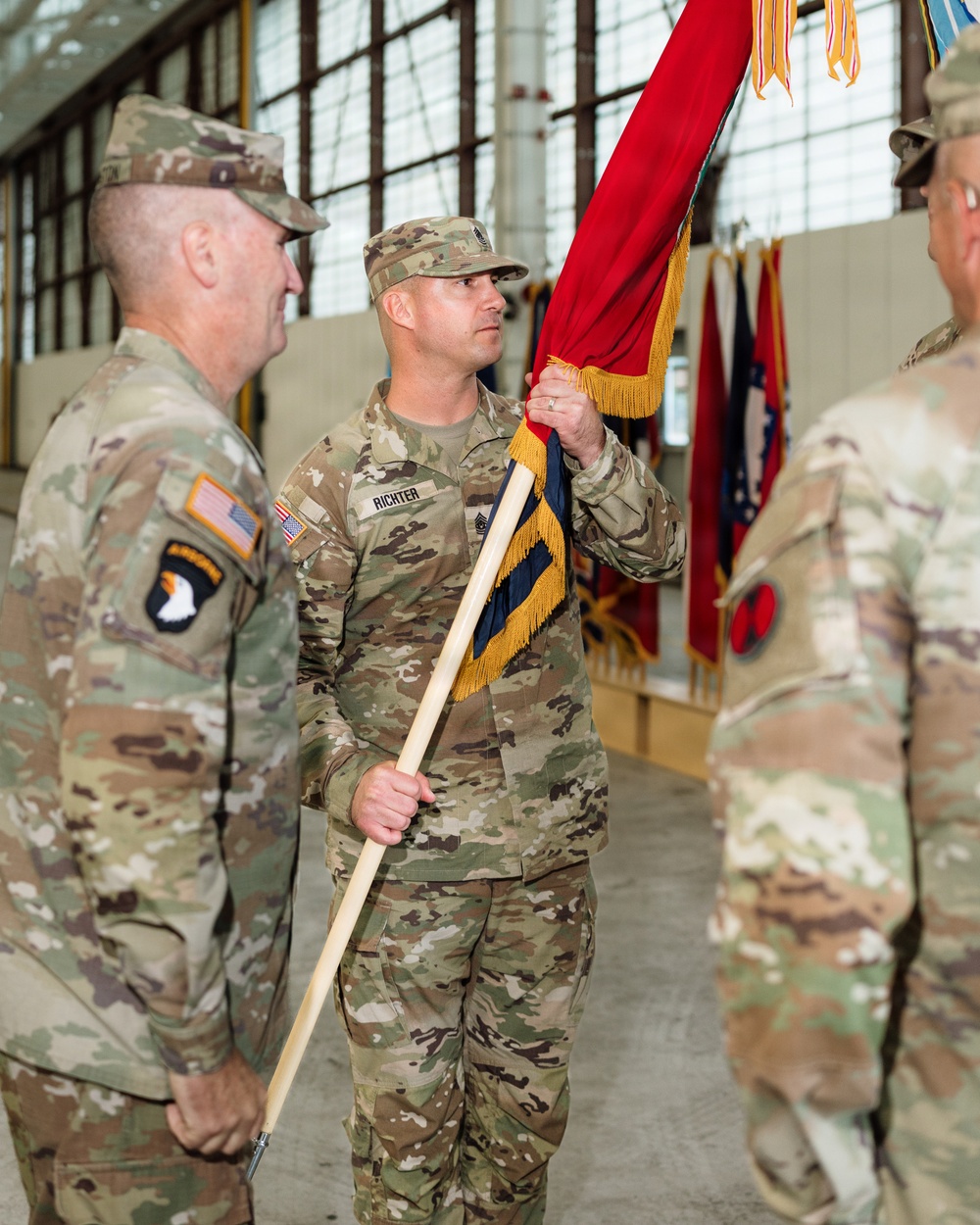 A U.S. Army military ceremony, likely a change of command or activation, taking place inside a hangar or armory. A soldier named RICHTER is holding a red guidon with gold fringe, flanked by other soldiers in OCP (Operational Camouflage Pattern) uniforms. Multiple unit colors and flags are visible in the background.