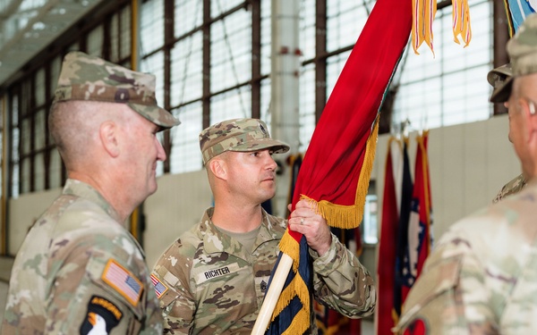 A U.S. Army military ceremony, likely a change of command or activation, taking place inside a hangar or armory. A soldier named RICHTER is holding a red guidon with gold fringe, flanked by other soldiers in OCP (Operational Camouflage Pattern) uniforms. Multiple unit colors and flags are visible in the background.