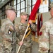 A U.S. Army military ceremony, likely a change of command or activation, taking place inside a hangar or armory. A soldier named RICHTER is holding a red guidon with gold fringe, flanked by other soldiers in OCP (Operational Camouflage Pattern) uniforms. Multiple unit colors and flags are visible in the background.