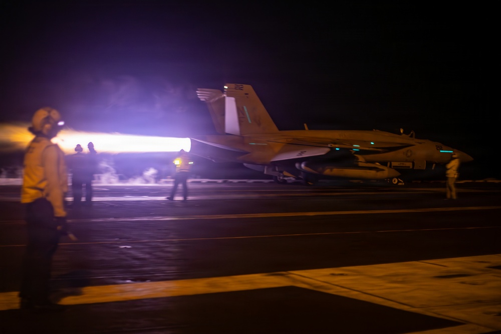 A night operations scene on an aircraft carrier flight deck showing a military jet aircraft with afterburners ignited during launch preparation. Deck crew in yellow jerseys are visible around the aircraft amid steam and exhaust. The intense engine exhaust flame illuminates the otherwise dark flight deck environment.