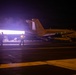 A night operations scene on an aircraft carrier flight deck showing a military jet aircraft with afterburners ignited during launch preparation. Deck crew in yellow jerseys are visible around the aircraft amid steam and exhaust. The intense engine exhaust flame illuminates the otherwise dark flight deck environment.