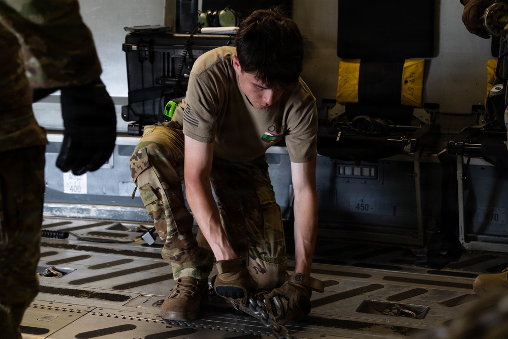 A military service member in OCP (Operational Camouflage Pattern) uniform is crouched down on the floor of what appears to be a military cargo aircraft, working with equipment or securing items to the aircraft floor. The individual is wearing a tan t-shirt over their uniform and appears to be manipulating tie-down fittings or cargo restraints on the aircraft's roller/tie-down floor system. Additional personnel and military equipment cases are visible in the background.