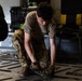 A military service member in OCP (Operational Camouflage Pattern) uniform is crouched down on the floor of what appears to be a military cargo aircraft, working with equipment or securing items to the aircraft floor. The individual is wearing a tan t-shirt over their uniform and appears to be manipulating tie-down fittings or cargo restraints on the aircraft's roller/tie-down floor system. Additional personnel and military equipment cases are visible in the background.