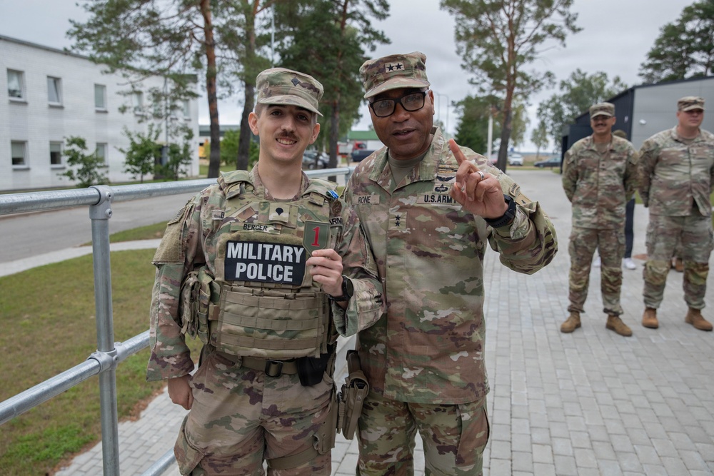Two U.S. Army soldiers pose together for a photo at what appears to be a military installation. The soldier on the left wears a 'Military Police' vest and a name tape reading 'BERGER,' while the taller soldier on the right displays two-star general rank insignia and a 'U.S. ARMY' tape, raising one finger. Two additional soldiers stand in the background near a building.