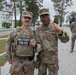 Two U.S. Army soldiers pose together for a photo at what appears to be a military installation. The soldier on the left wears a 'Military Police' vest and a name tape reading 'BERGER,' while the taller soldier on the right displays two-star general rank insignia and a 'U.S. ARMY' tape, raising one finger. Two additional soldiers stand in the background near a building.