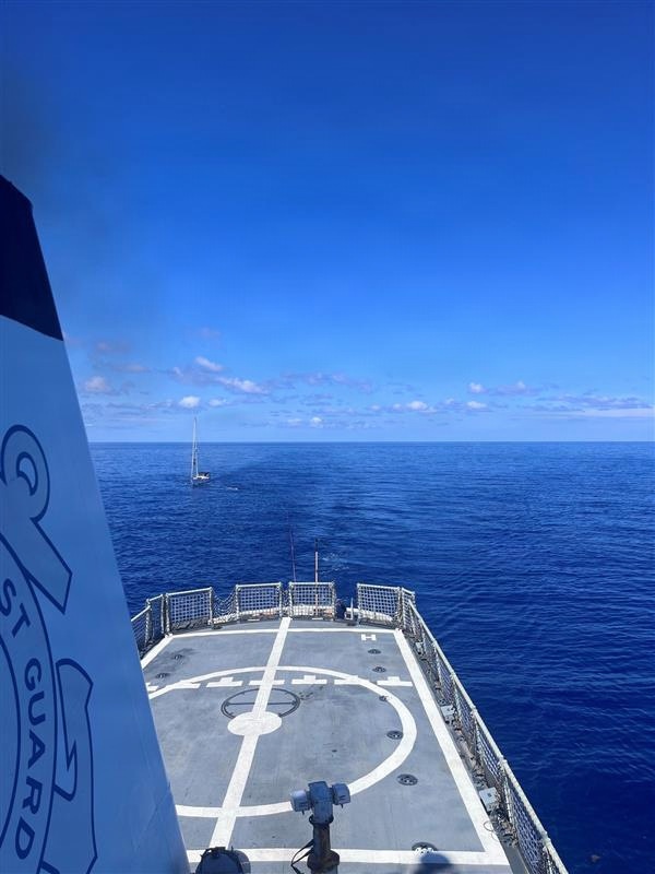 The image is taken from the elevated deck of a U.S. Coast Guard vessel, looking forward over a helicopter landing pad (helipad) toward the open ocean. A small sailboat is visible in the distance, suggesting a possible intercept, patrol, or rescue operation. The weather is clear with calm seas and bright blue skies.