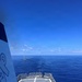 The image is taken from the elevated deck of a U.S. Coast Guard vessel, looking forward over a helicopter landing pad (helipad) toward the open ocean. A small sailboat is visible in the distance, suggesting a possible intercept, patrol, or rescue operation. The weather is clear with calm seas and bright blue skies.