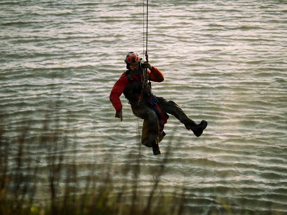 A rescue or search-and-rescue technician is suspended from a cable or rope over a body of water, likely being lowered or hoisted by a helicopter or crane. The individual is wearing full safety gear including a helmet, harness, and protective clothing. The water below appears choppy, suggesting outdoor conditions that may be part of a real operation or training exercise.