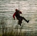 A rescue or search-and-rescue technician is suspended from a cable or rope over a body of water, likely being lowered or hoisted by a helicopter or crane. The individual is wearing full safety gear including a helmet, harness, and protective clothing. The water below appears choppy, suggesting outdoor conditions that may be part of a real operation or training exercise.