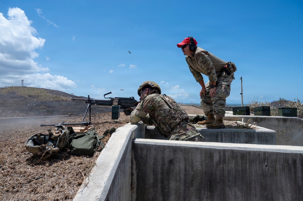 Two military personnel in OCP (Operational Camouflage Pattern) uniforms are conducting a machine gun firing exercise at an outdoor range. One soldier is in a prone/kneeling position operating what appears to be an M249 or M240 light machine gun, while a second individual wearing a red cap acts as a range safety officer or instructor observing the shooter. A spent casing is visibly ejecting from the weapon, indicating active firing.