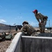 Two military personnel in OCP (Operational Camouflage Pattern) uniforms are conducting a machine gun firing exercise at an outdoor range. One soldier is in a prone/kneeling position operating what appears to be an M249 or M240 light machine gun, while a second individual wearing a red cap acts as a range safety officer or instructor observing the shooter. A spent casing is visibly ejecting from the weapon, indicating active firing.