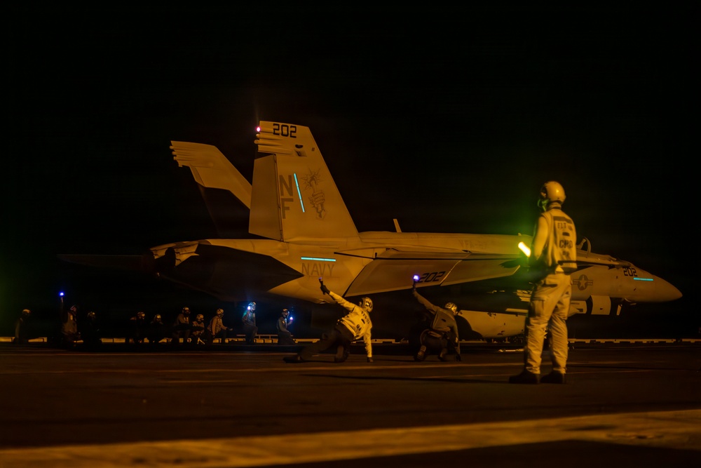 A U.S. Navy F/A-18 Hornet or Super Hornet (bureau number 202, tail code 'NF') is being prepared for a night launch on an aircraft carrier flight deck. Multiple flight deck crew members in various colored jerseys are visible performing pre-launch duties around the aircraft. The scene is illuminated by a combination of aircraft lighting, deck lighting, and ambient orange glow, consistent with nighttime carrier flight operations.