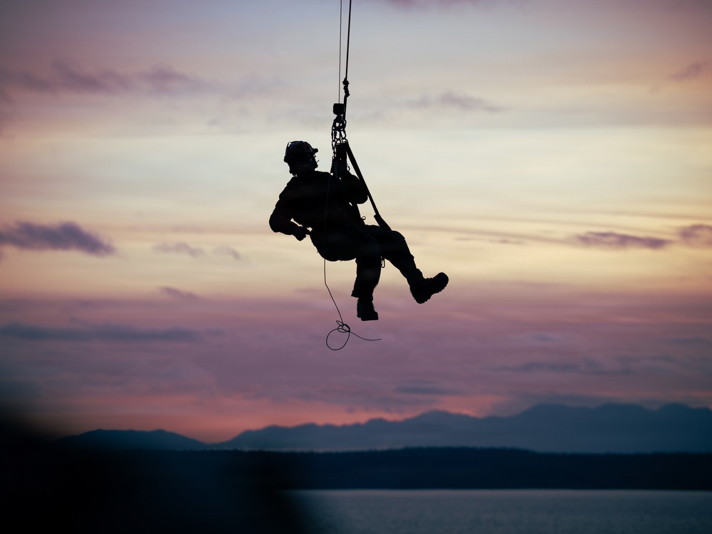 A silhouetted figure is suspended from a rope or cable against a dramatic sunset sky, appearing to be engaged in a military or rescue hoist operation. The person is equipped with tactical gear including a helmet and harness, dangling over a body of water with mountain ridgelines visible in the background. The image is captured in strong silhouette contrast against warm pink, orange, and purple twilight tones.