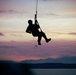 A silhouetted figure is suspended from a rope or cable against a dramatic sunset sky, appearing to be engaged in a military or rescue hoist operation. The person is equipped with tactical gear including a helmet and harness, dangling over a body of water with mountain ridgelines visible in the background. The image is captured in strong silhouette contrast against warm pink, orange, and purple twilight tones.