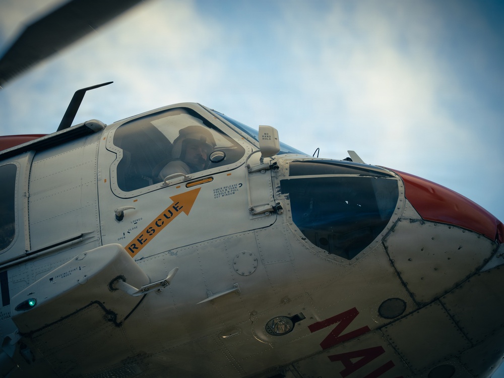 Close-up photograph of a rescue helicopter cockpit area, showing the nose and pilot's canopy section. The aircraft features a white and red color scheme with a prominent orange 'RESCUE' label and directional arrow on the fuselage. A pilot or crew member is partially visible through the cockpit glass.