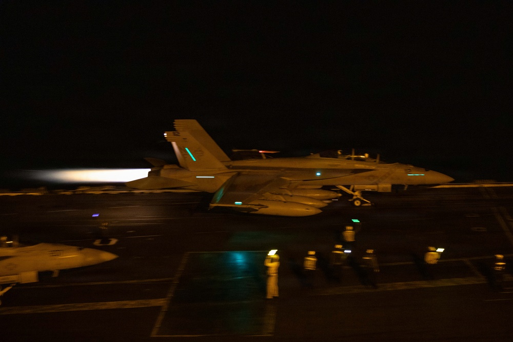 A military jet aircraft, consistent with an F/A-18 Hornet or Super Hornet, is captured during a night launch from what appears to be an aircraft carrier flight deck. The aircraft's engines are producing visible afterburner exhaust glow on the left side of the frame. Deck crew personnel and flight deck lighting are visible in the foreground and background.