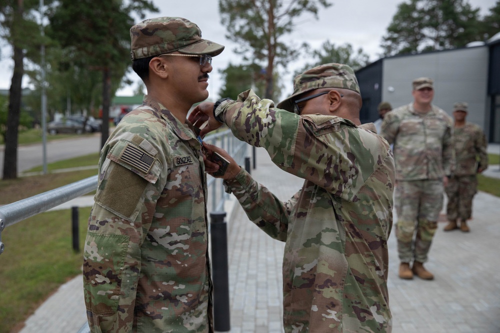Two U.S. Army soldiers in OCP (Operational Camouflage Pattern) uniforms are engaged in what appears to be a pinning ceremony, with one soldier attaching something to the collar or chest area of the other. The soldier receiving the pin has a name tape reading 'GONZALEZ' and wears a U.S. flag patch on the right shoulder. Additional soldiers are visible in the background in a paved outdoor area surrounded by trees.