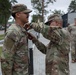 Two U.S. Army soldiers in OCP (Operational Camouflage Pattern) uniforms are engaged in what appears to be a pinning ceremony, with one soldier attaching something to the collar or chest area of the other. The soldier receiving the pin has a name tape reading 'GONZALEZ' and wears a U.S. flag patch on the right shoulder. Additional soldiers are visible in the background in a paved outdoor area surrounded by trees.
