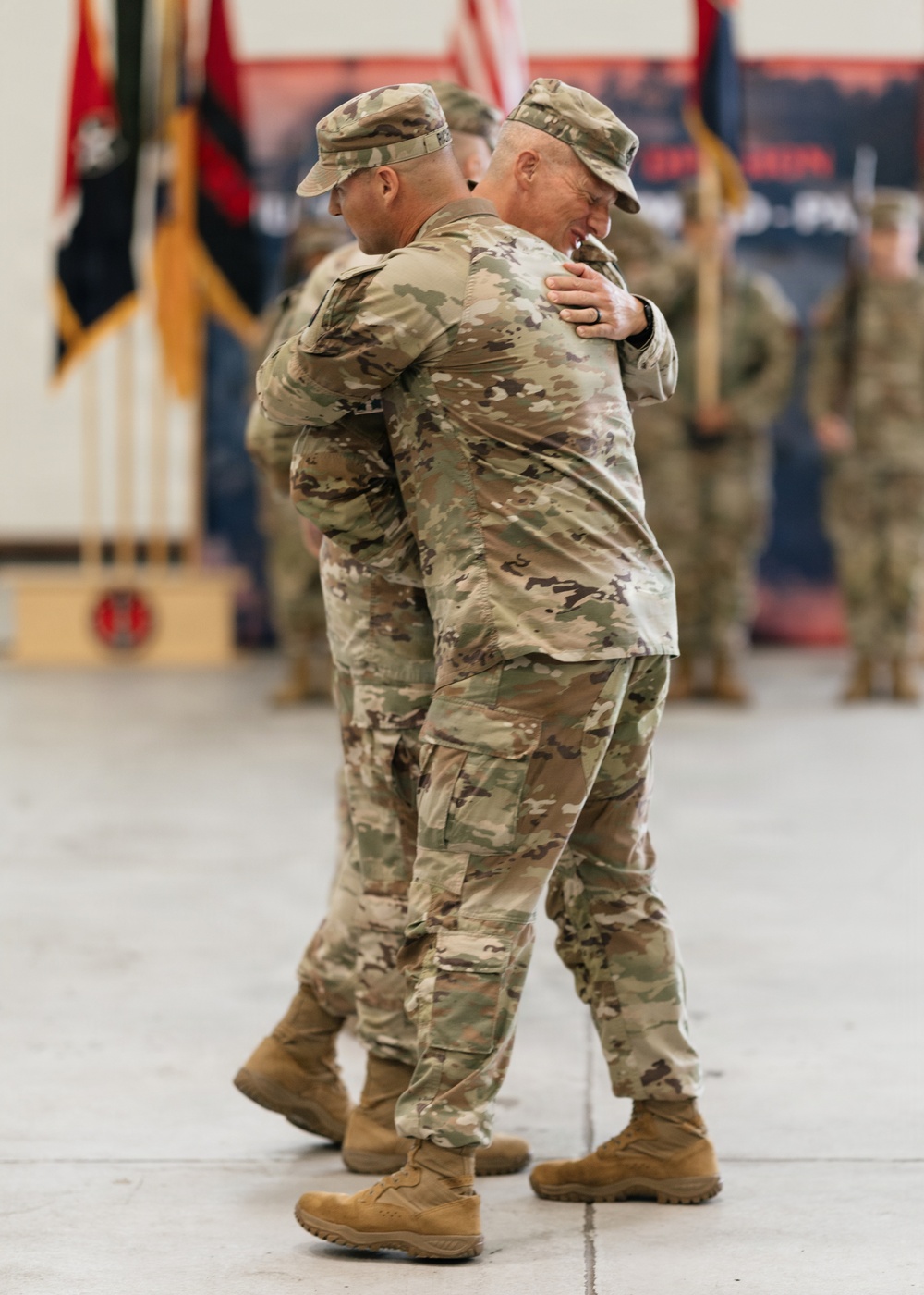 Two U.S. Army soldiers in Operational Camouflage Pattern (OCP) uniforms are embracing during what appears to be a formal military ceremony, likely a change of command or redeployment event. The setting is an indoor military facility such as a hangar or gymnasium, with unit guidons, flags, and formation soldiers visible in the background. The embrace suggests a moment of personal significance, such as a farewell or welcome.