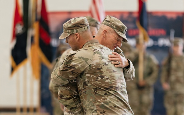 Two U.S. Army soldiers in Operational Camouflage Pattern (OCP) uniforms are embracing during what appears to be a formal military ceremony, likely a change of command or redeployment event. The setting is an indoor military facility such as a hangar or gymnasium, with unit guidons, flags, and formation soldiers visible in the background. The embrace suggests a moment of personal significance, such as a farewell or welcome.