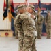 Two U.S. Army soldiers in Operational Camouflage Pattern (OCP) uniforms are embracing during what appears to be a formal military ceremony, likely a change of command or redeployment event. The setting is an indoor military facility such as a hangar or gymnasium, with unit guidons, flags, and formation soldiers visible in the background. The embrace suggests a moment of personal significance, such as a farewell or welcome.