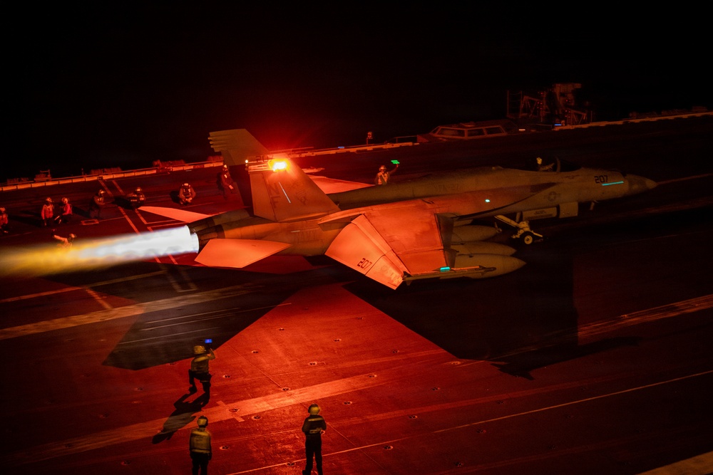 A night carrier operations scene showing an F/A-18 Hornet or Super Hornet launching from an aircraft carrier deck with afterburners fully engaged, bathing the flight deck in intense orange-red light. A second aircraft marked '207' is visible in the background awaiting its turn. Multiple flight deck crew members in safety gear are visible at their stations.
