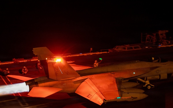 A night carrier operations scene showing an F/A-18 Hornet or Super Hornet launching from an aircraft carrier deck with afterburners fully engaged, bathing the flight deck in intense orange-red light. A second aircraft marked '207' is visible in the background awaiting its turn. Multiple flight deck crew members in safety gear are visible at their stations.
