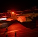 A night carrier operations scene showing an F/A-18 Hornet or Super Hornet launching from an aircraft carrier deck with afterburners fully engaged, bathing the flight deck in intense orange-red light. A second aircraft marked '207' is visible in the background awaiting its turn. Multiple flight deck crew members in safety gear are visible at their stations.