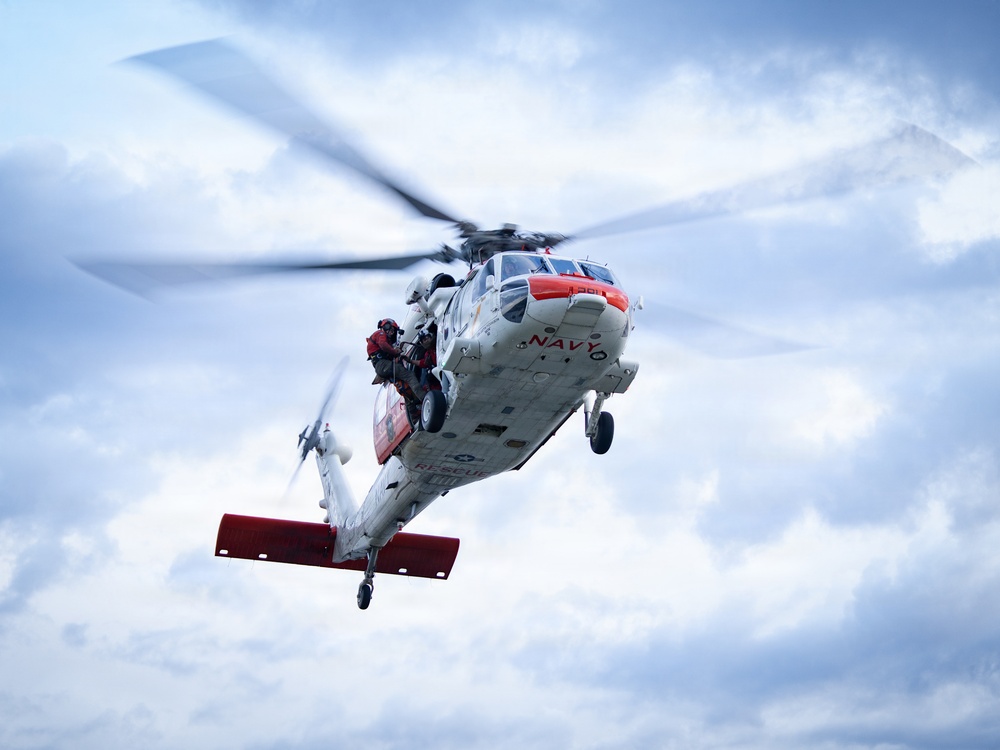 A U.S. Navy search and rescue helicopter, marked 'NAVY' and 'RESCUE' in red lettering, is captured in flight against a partly cloudy sky. The white and orange-accented helicopter has a crew member in red gear visible at the open side door. The image appears to depict an active rescue operation or training exercise.