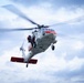 A U.S. Navy search and rescue helicopter, marked 'NAVY' and 'RESCUE' in red lettering, is captured in flight against a partly cloudy sky. The white and orange-accented helicopter has a crew member in red gear visible at the open side door. The image appears to depict an active rescue operation or training exercise.