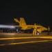 A U.S. Navy aircraft carrier flight deck at night showing a jet aircraft preparing for or undergoing catapult launch operations. The aircraft, numbered 504 with tail markings 'NF', has its engines at high thrust producing visible exhaust plumes. Multiple flight deck personnel in yellow jerseys are visible observing and managing the launch sequence.